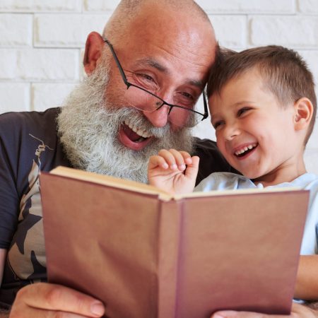 Bearded aged man and his grandson are having fun reading a book together while sitting in cozy armchair at home