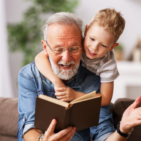 Happy family elderly  man and little boy smiling r while sitting on couch and reading fascinating fairy tale together at home