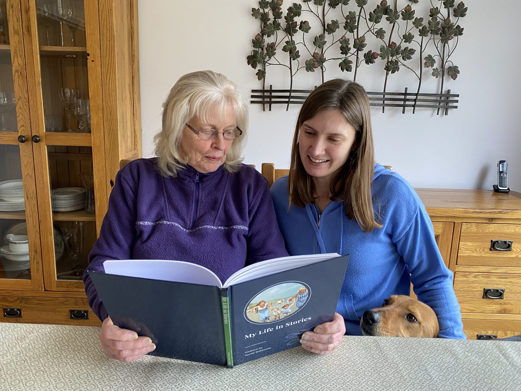 A mother and daughter reading her YourStory life story book together at the kitchen table, with the family dog looking on
